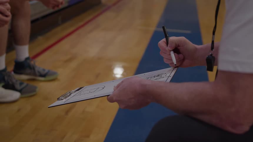 A coach is filmed in a closeup lowangle shot as he kneel and use a dark pen to draw on a clipboard showing a basketball court diagram while the lower legs and athletic shoes of several other people stand on the wooden court floor behind him