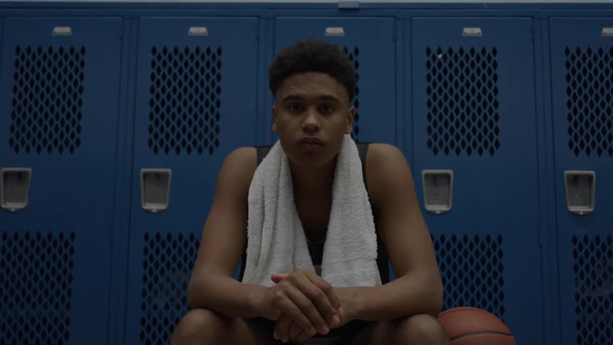 A young basketball player with a towel around his neck sits in front of blue lockers staring intently with a ball at his side