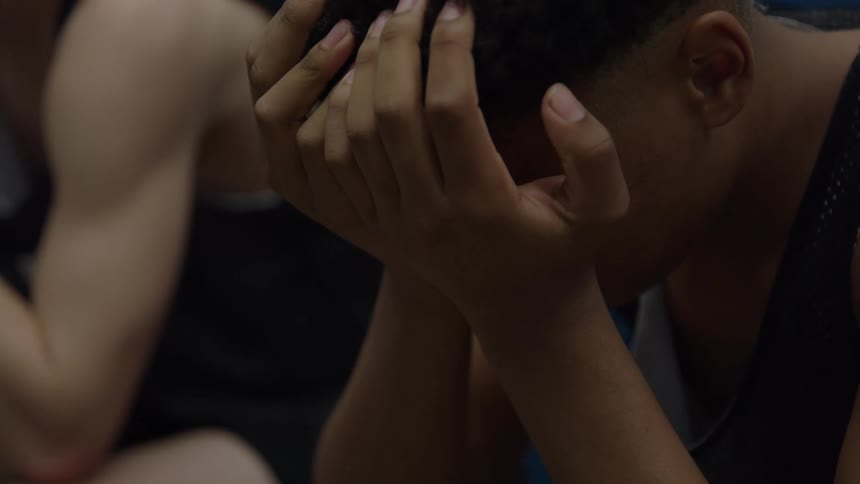 A very close view shows a young man with curly hair and a black tank top sitting dejectedly with his hands covering his face in a dimly lit locker room