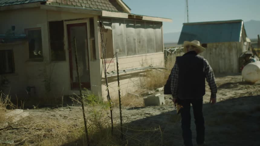 A man in a hat carries a gun toward a house