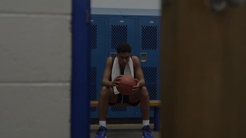 A medium straight on view through a doorway shows a young man sitting on a wooden bench holding a basketball and a white towel in a blue locker room