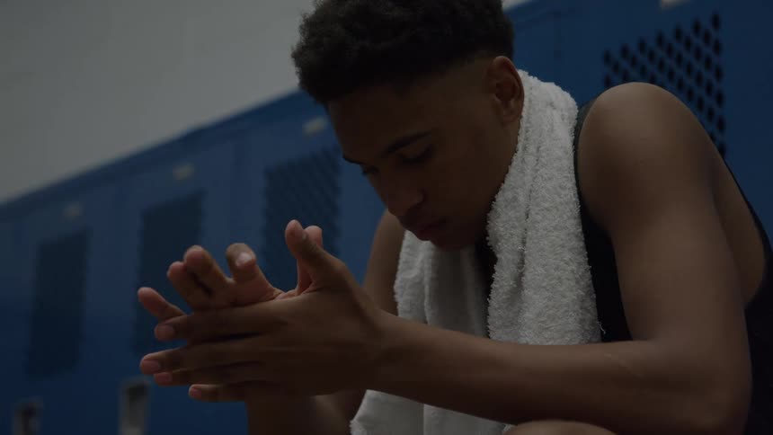 A young man in a black tank top with a white towel draped around his neck sits in a dark locker room with blue lockers rubbing his hands together and looking down with a somber or contemplative expression