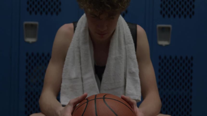 A very close low angle view shows a young man with curly brown hair sitting on a wooden bench holding a basketball and a white towel around his neck looking up with a determined expression against a backdrop of blue lockers