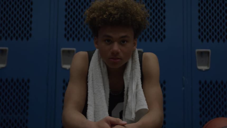 A young man with curly hair sits hunched over with his hands clasped wearing a dark sleeveless jersey and a white towel draped around his neck in front of a wall of dark blue metal lockers with a basketball visible on the right