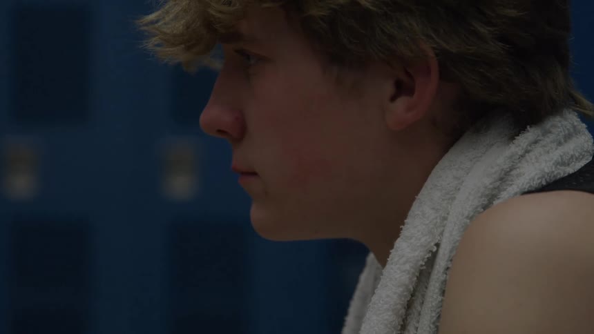 A profile close view of a young man with blonde hair and a white towel over his shoulder looks down and concentrates in a dimly lit blue locker room or gymnasium