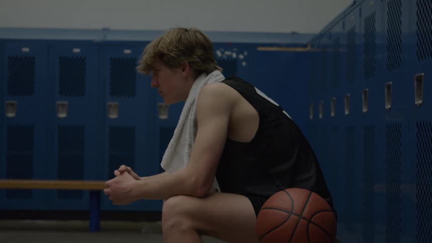 A teenage boy wearing a dark sleeveless jersey and shorts sits on a wooden bench in a locker room with a basketball on his side and a towel in his neck