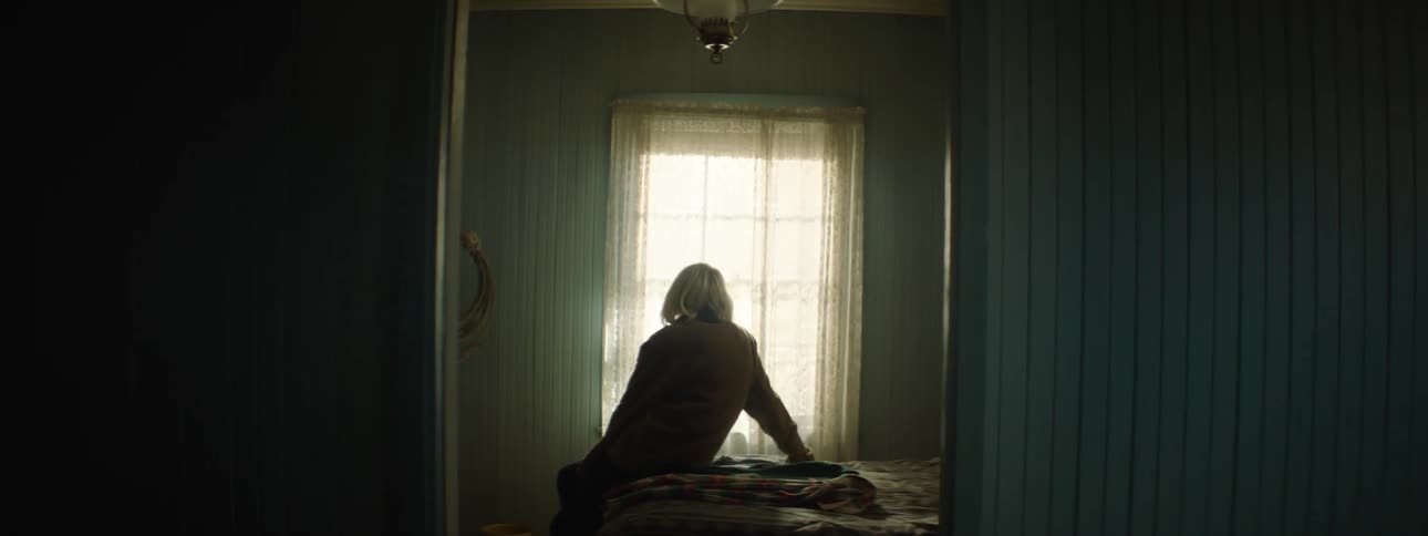 An old woman is sitting on a bed staring toward a window