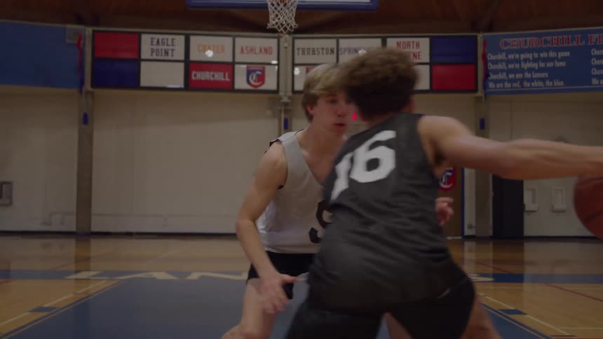 Two male youth basketball players one wearing a dark jersey with the number 16 and the other wearing a light jersey with the number 9 engage in a oneonone drill on a wooden court floor