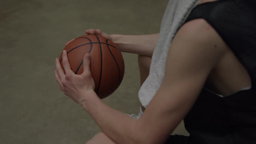 A basketball player in a black jersey sits holding a ball with both hands a towel draped over his shoulder