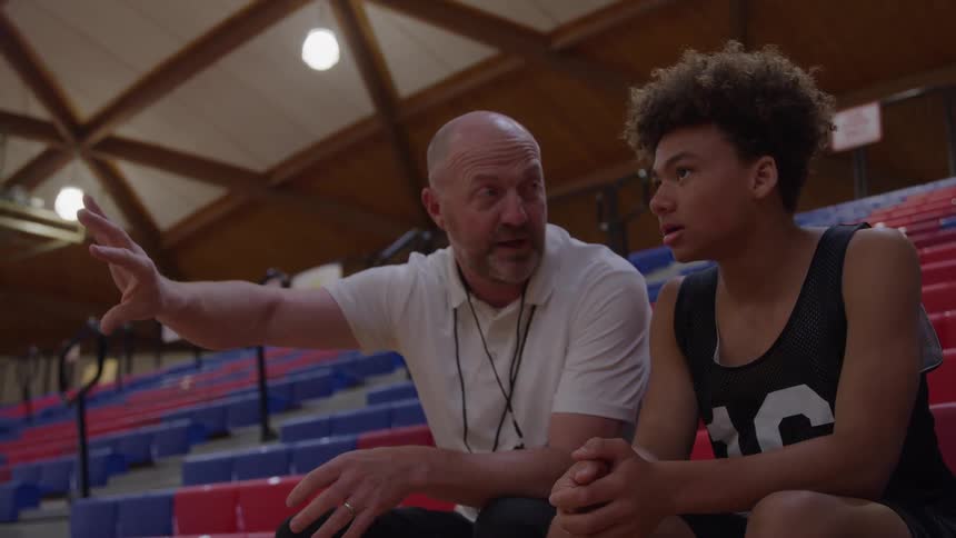 A bald coach in a white shirt and a young man in a black basketball jersey sit on red and blue bleachers in a dimly lit gymnasium having a serious discussion as the coach gestures emphatically