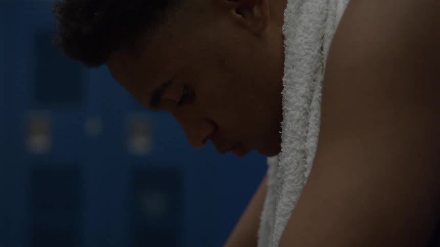 A young man with a white towel around his neck looks down in a blue locker room appearing thoughtful and sad