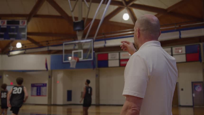A bald man wearing a white shirt stands with his back mostly to the camera on a wooden court watching and pointing at several young male basketball players who are moving and shooting a ball in the distance in an indoor gymnasium