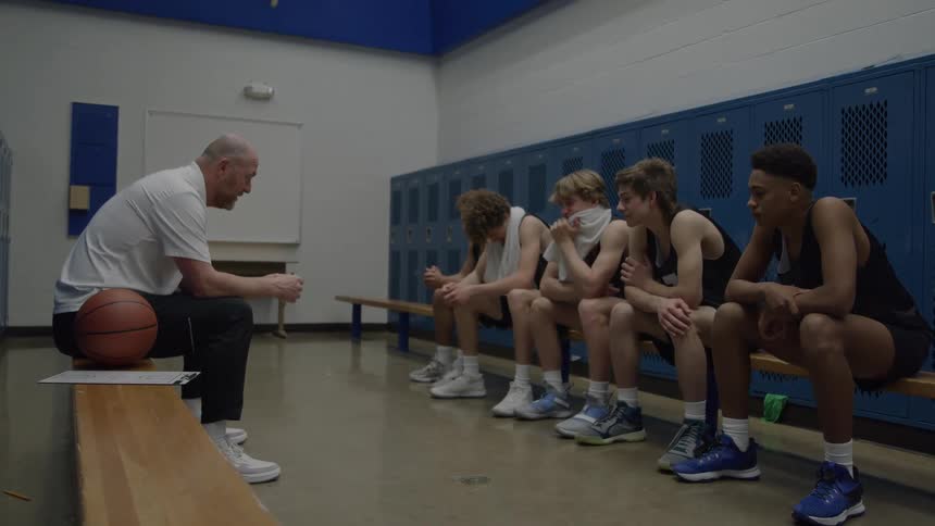 A medium view of a bald coach in a white shirt sitting on a stool and talking to five young male basketball players sitting dejectedly on a bench against blue lockers in a locker room with a basketball nearby