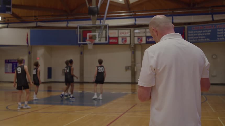A medium rear view of a bald coach in a white shirt watching and holding a clipboard as a group of four young male basketball players in black jerseys practice on the court under the hoop