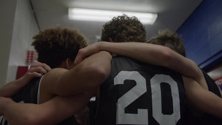 A group of basketball players in black jerseys huddle with arms around each other under a bright locker room light