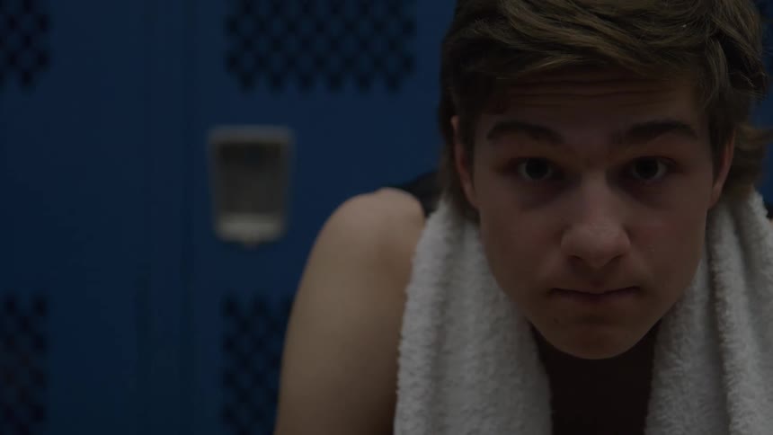 A young man with a white towel around his neck sits in a locker room looking straight ahead with a focused slightly tense expression