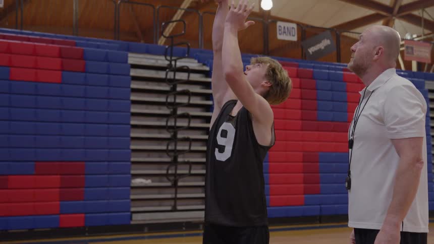 A middleaged man and a young male athlete stand on a wood gymnasium floor with the man demonstrating and instructing the youth on the motion of throwing an orange basketball while the youth practices the same movement under the mans observation