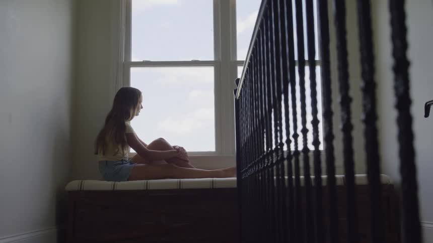 A side view shows a young woman with long hair wearing a tank top and shorts sitting alone on a window seat and looking out a bright window next to a dark metal railing