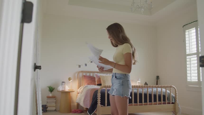 A side view of a young woman with long hair wearing a yellow shirt and denim shorts stands in her bright bedroom looking at some papers in her hands near a wooden bed frame and a shuttered window