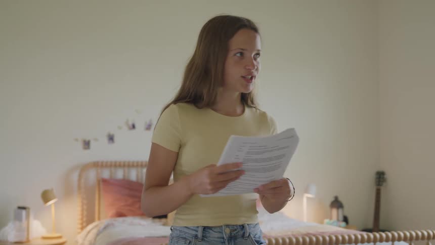 Teen aged female speaks out loud while reading from a document as she walks around in her bedroom