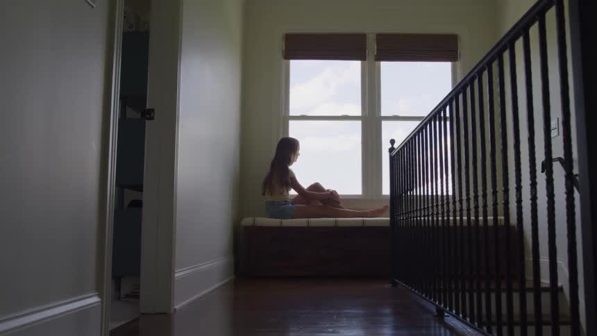 A young female sits on a padded bench at the end of a hallway near a window she seems to be in deep thought