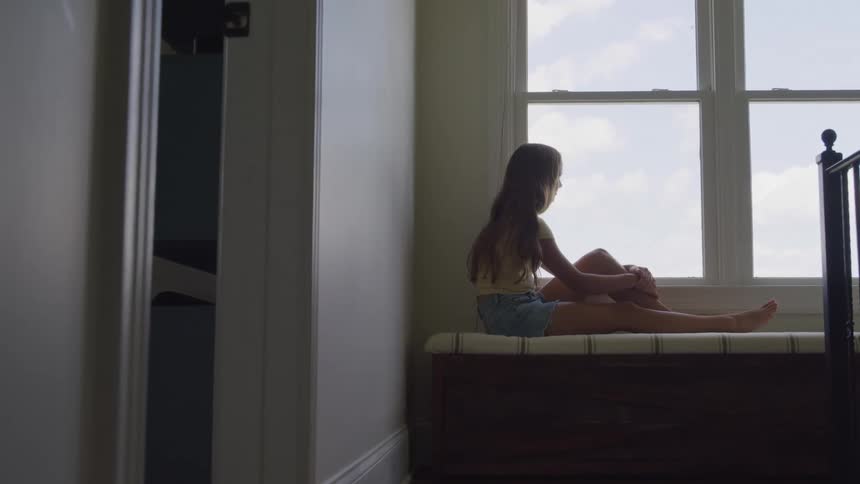 A wide interior view shows a young woman sitting quietly on a dark window seat bench wearing a pale yellow top and denim shorts while looking out a bright window at the sky