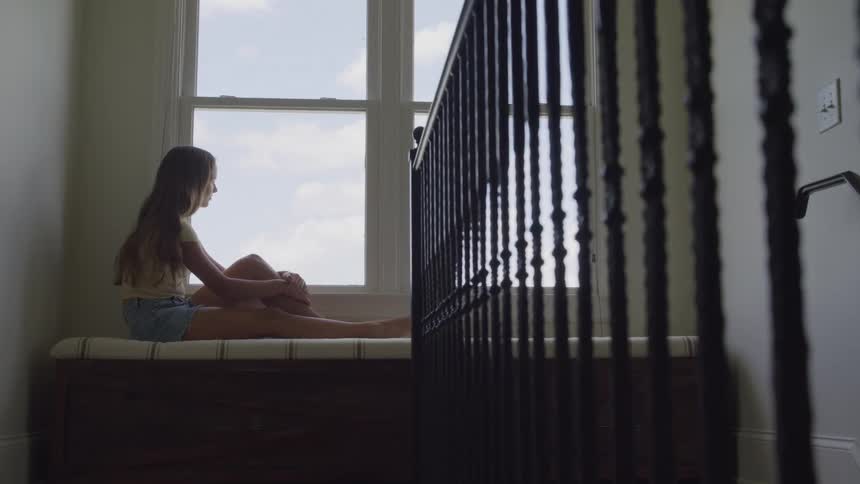 A teenage girl sits on a bed beside a window gazing outside at midday as sunlight fills the room