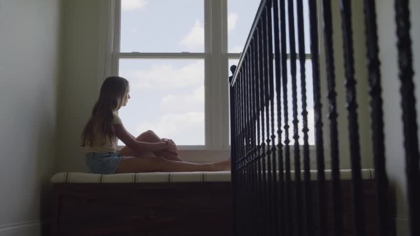 A girl sits on a bench gazing out through a bright indoor window