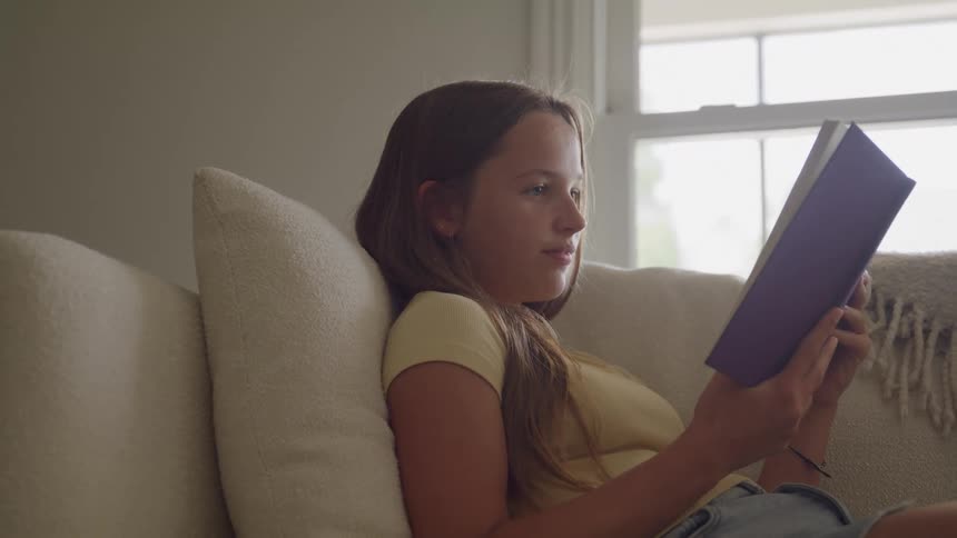 A young female rests back on a couch as she read a book in a living room