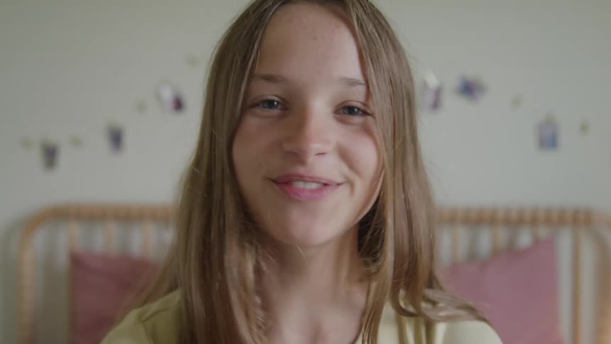A teenage girl records a selfie video in her bedroom at midday smiling brightly as sunlight fills the space