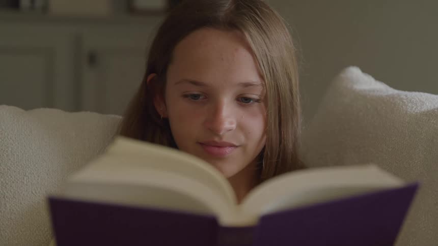 A young girl relaxes on a couch reading a purple book with a faint content smile