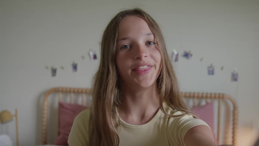 A young female with long hair holds a device toward herself in an indoor room with a bed and rattan headboard visible in the background