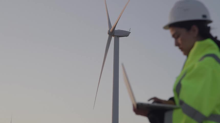 A young woman in a green vest and white hard hat operates a laptop adjusting the speed of a wind turbine at a windmill site