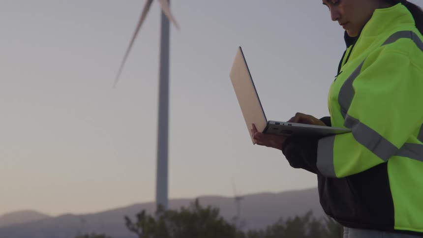 A female technician works on a laptop in front of a windmill turbine at dusk