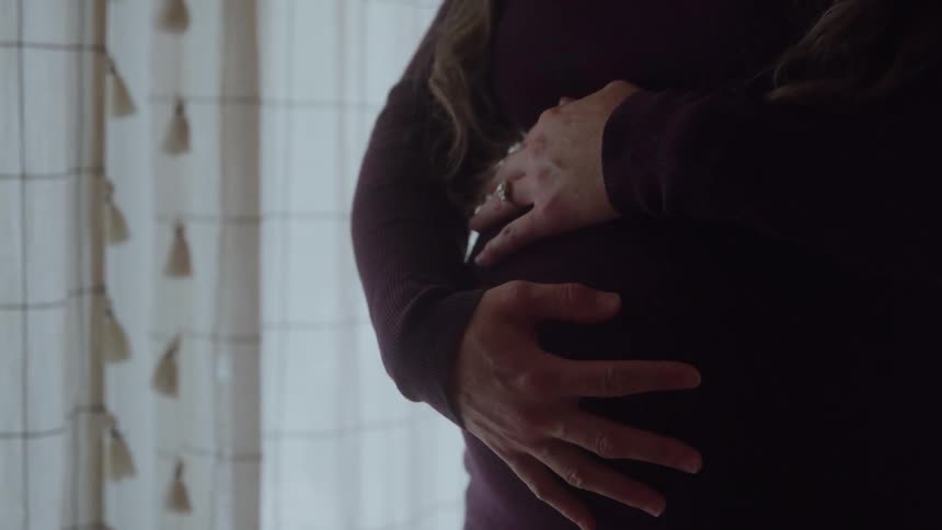 A pregnant woman in a maroon dress cradles her belly with both hands near a window with tassel curtains