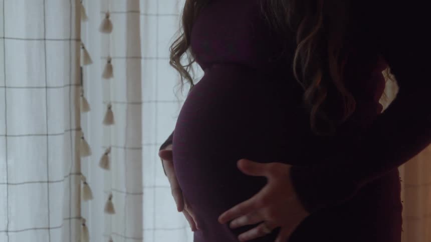Expectant mother standing near the window strokes her abdomen
