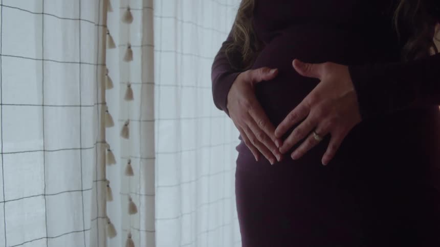 A pregnant woman in a maroon dress cradles her belly with both hands near a softly lit window
