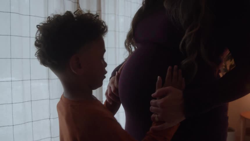 A pregnant woman stands indoors with her young son who gently plays with her baby bump sharing a tender moment