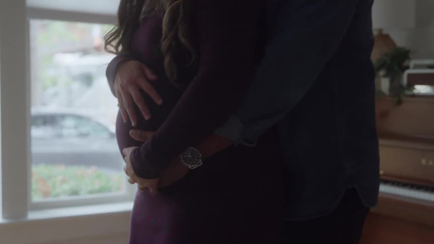 A close medium view shows a pregnant woman wearing a purple dress being embraced from behind by a man as they both place their hands protectively over her large belly while standing near a window indoors