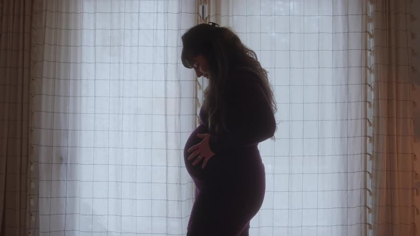 A medium profile silhouette of a pregnant woman wearing a dark dress as she lovingly cradles her large baby bump with her hands while standing in front of a bright window with sheer curtains