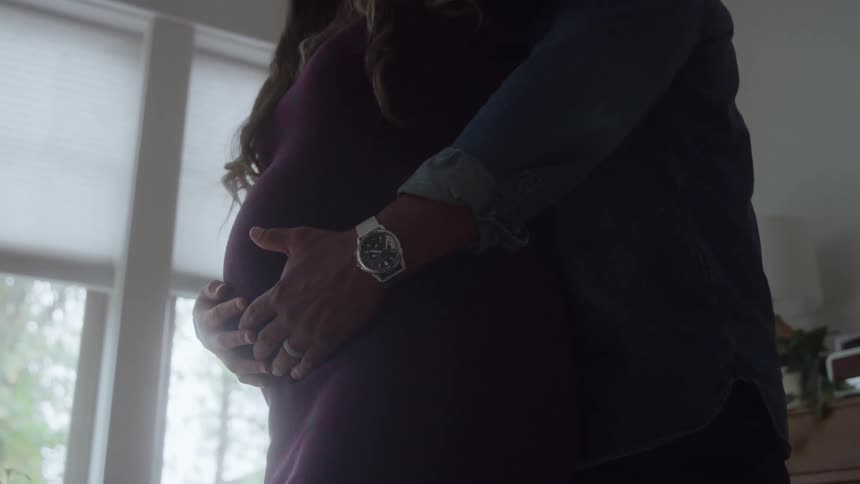Couple embraces as the man holds his pregnant partners belly by a window