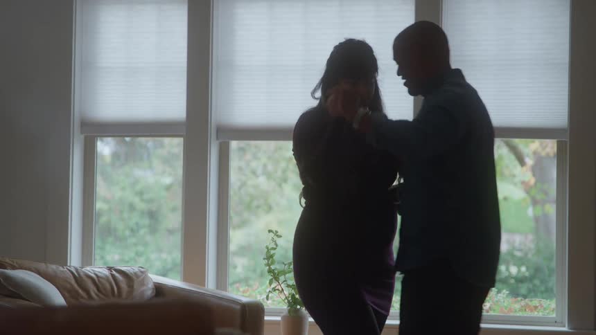 A pregnant woman dances with her male partner in the living room at midday sharing a romantic and playful moment together