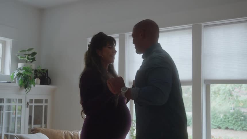 A pregnant woman dances with her male partner in the living room at midday sharing a romantic and playful moment together