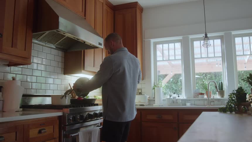 A man cooks in a sunlit kitchen pouring food from a pan into a pot on the stove beside wooden cabinets and large windows