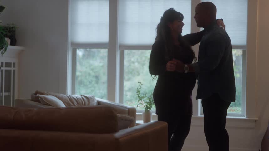 A smiling pregnant woman stands by bright windows as a man bends to her belly then they dance in a cozy living room