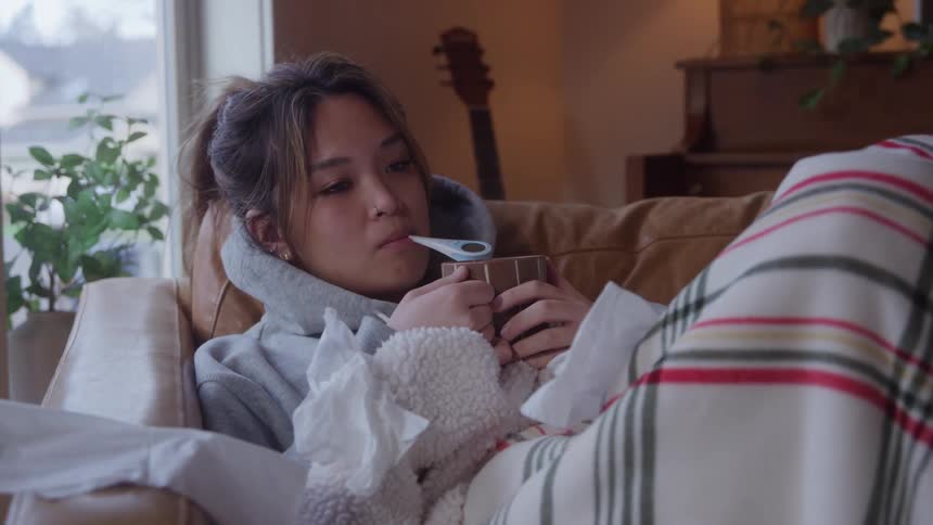 A sick woman rests on a couch with a thermometer in a cozy living room