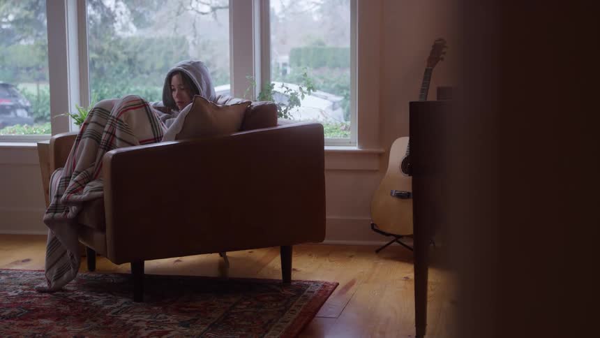 A medium cozy interior view shows a woman wearing a gray hoodie and wrapped in a plaid blanket sitting on a brown armchair and looking down at a book next to a piano and a guitar in front of a rainy window
