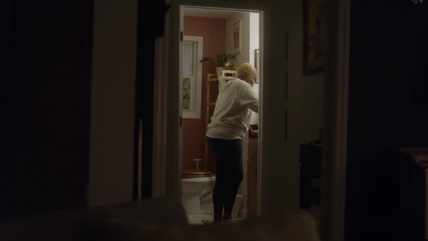 A woman stands inside a bathroom at night wearing a sweatshirt as the room glows with artificial light