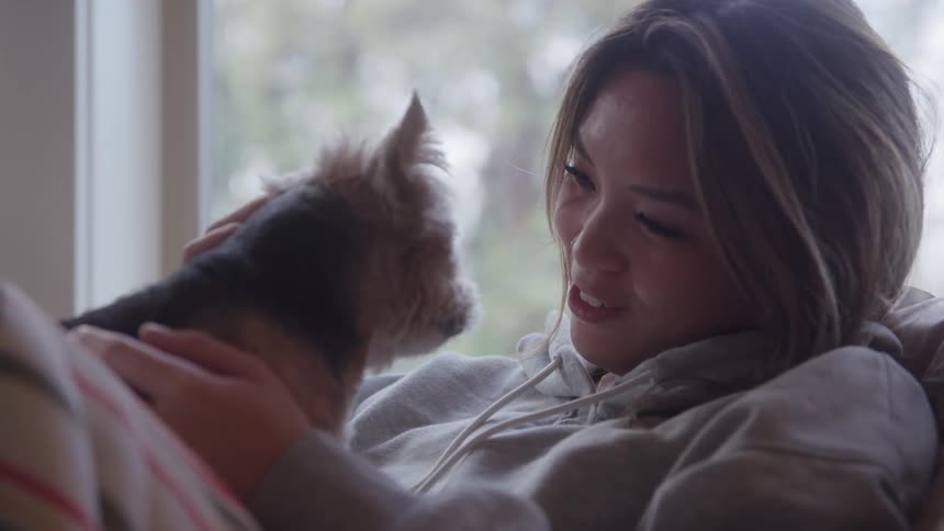 A woman wearing a grey hooded sweatshirt lies indoors near a window holding a small darkhaired dog close to her face and smiling gently while she uses one hand to stroke the animals chest