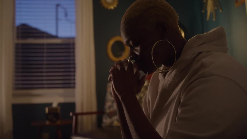 A woman with short blonde hair and large hoop earrings clasps her hands in a dim room appearing deep in thought or prayer